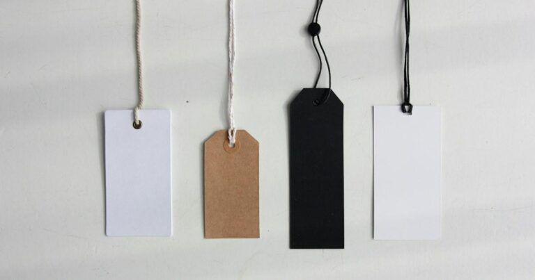Four blank paper tags in white, kraft brown and black hanging on strings against a pale grey wall